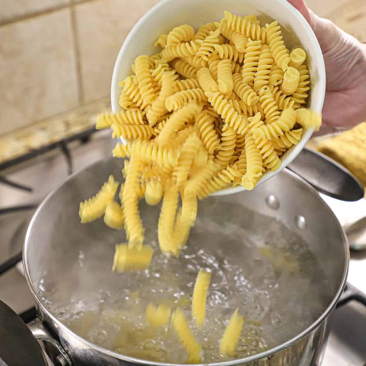 A person transferring uncooked fusilli pasta into a pot of boiling water on a gas stove.