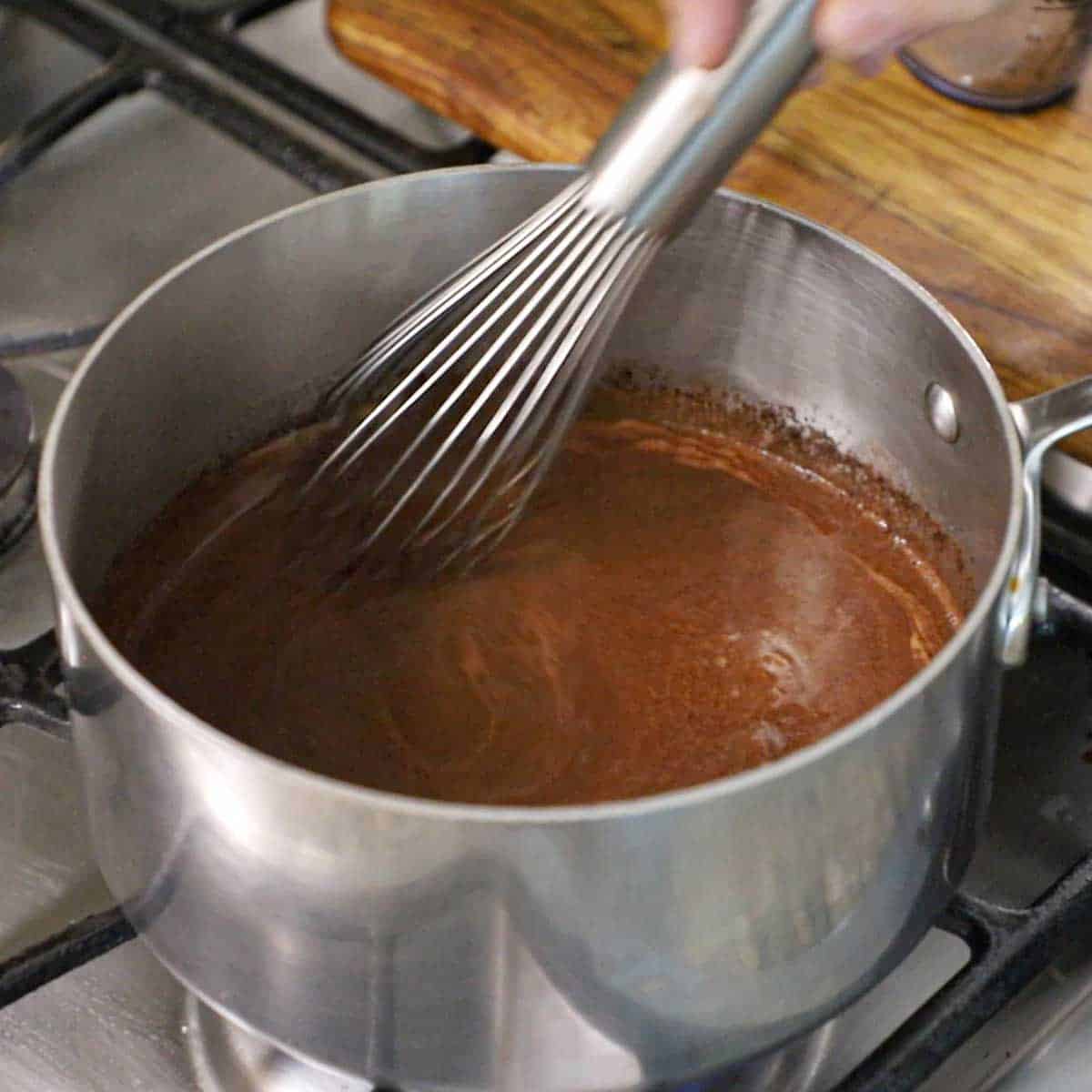 A person using a whisk to stir cocoa powder that is mixed with melted butter and brewed coffee in a saucepan on a gas stove.