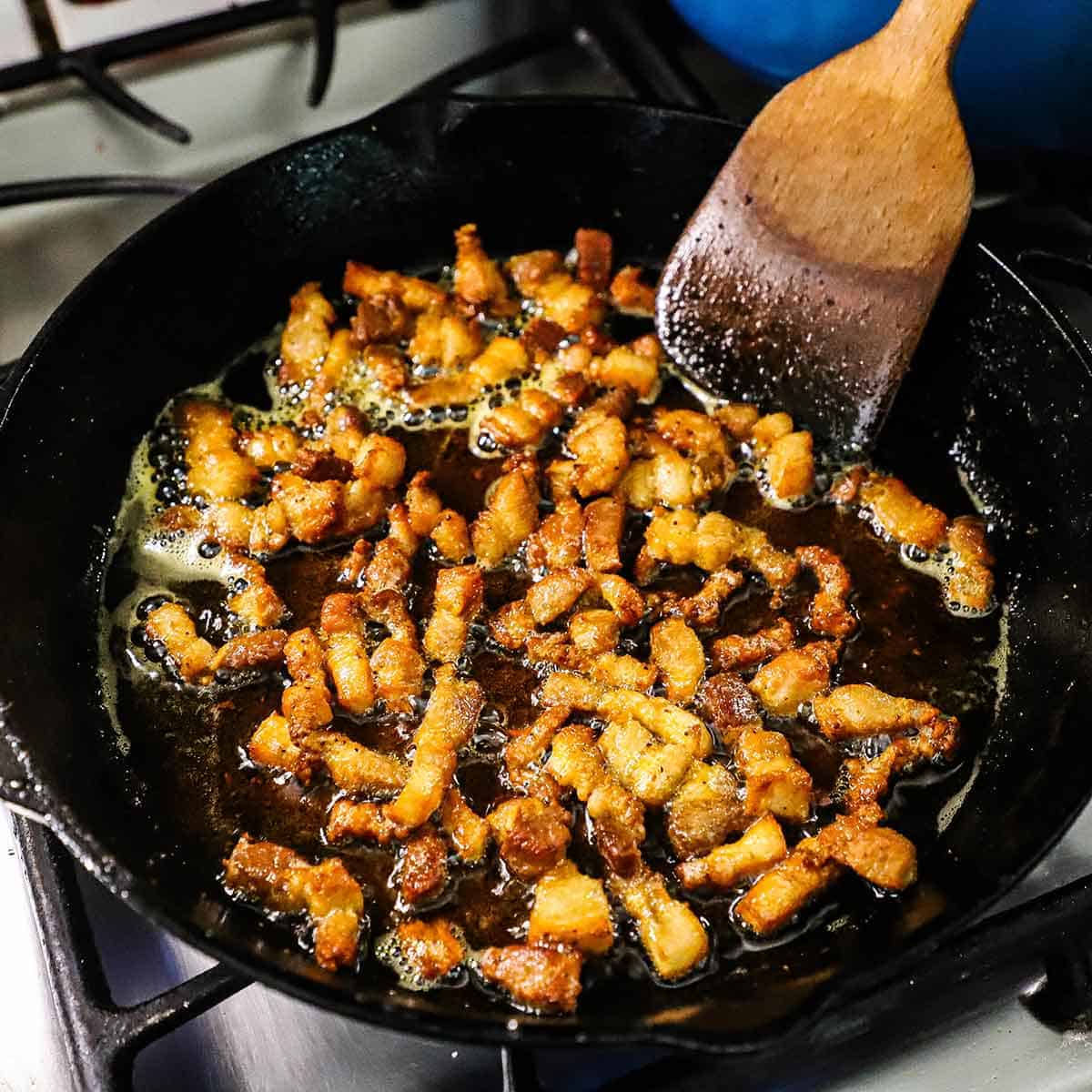 A person using a wooden spatula to stir crispy strips of pork belly being cooked in a large cast-iron skillet.