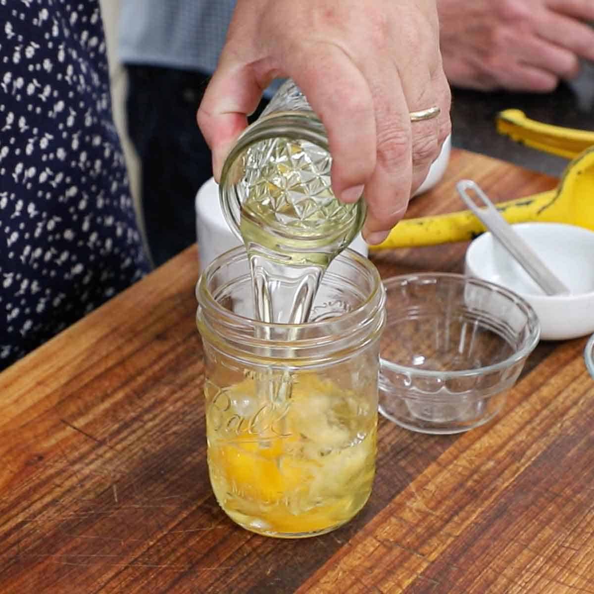 A person pouring olive oil from one jar into another jar that also contains a whole egg, an egg yolk, Dijon mustard, lemon juice, and white vinegar.