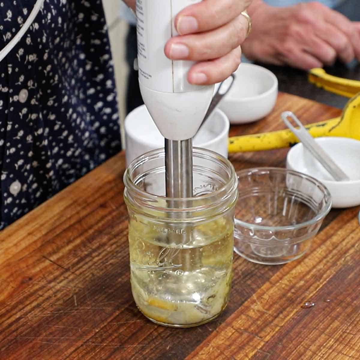 A person using an immersion blender to blend oil, an egg yolk, an egg, vinegar, and lemon juice in a glass jar on a wooden cutting board.