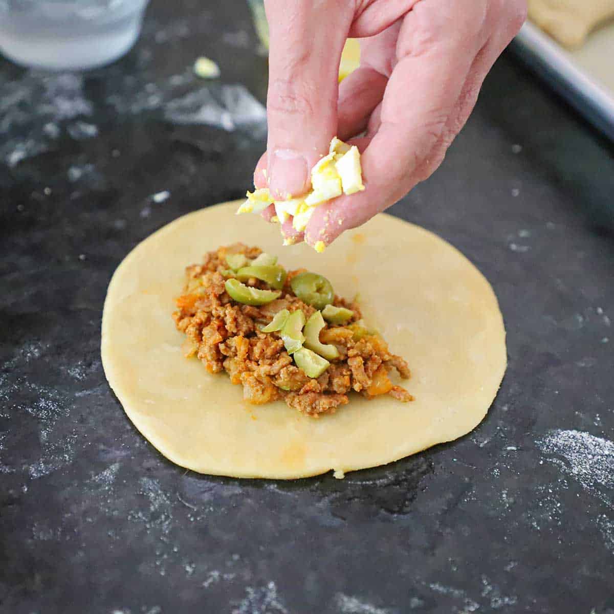 A person using his fingers to place chopped hard boiled eggs over a pile of beef filling that is the center of an empanada circular piece of dough.