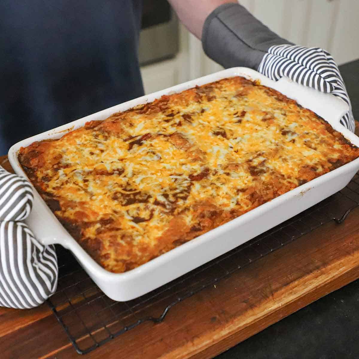 A person wearing oven mitts to place a freshly baked beef enchilada casserole in a baking dish onto a baking rack resting on a wooden cutting board.