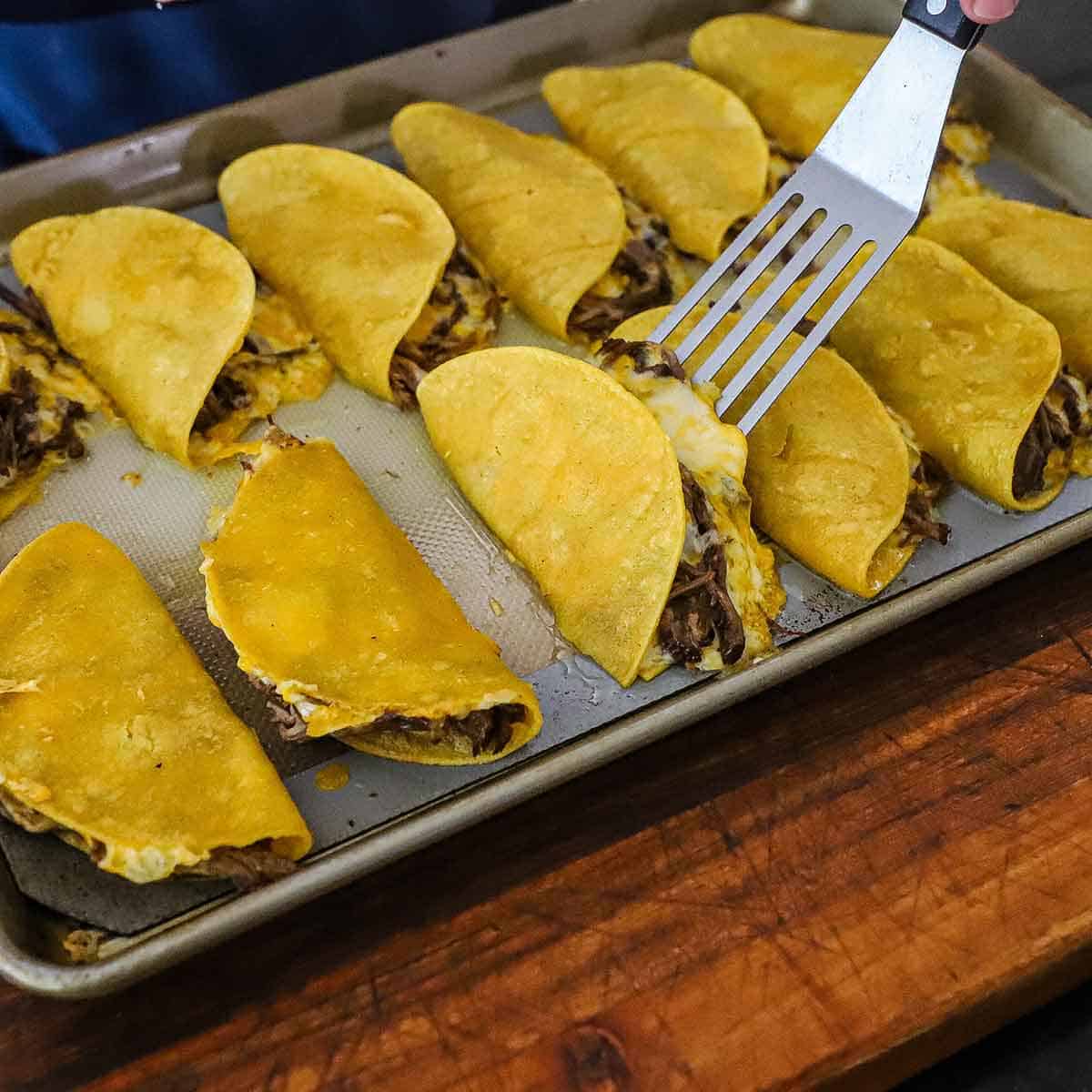 A person using a metal spatula to flip over a baked brisket taco on a baking pan filled with more brisket tacos.