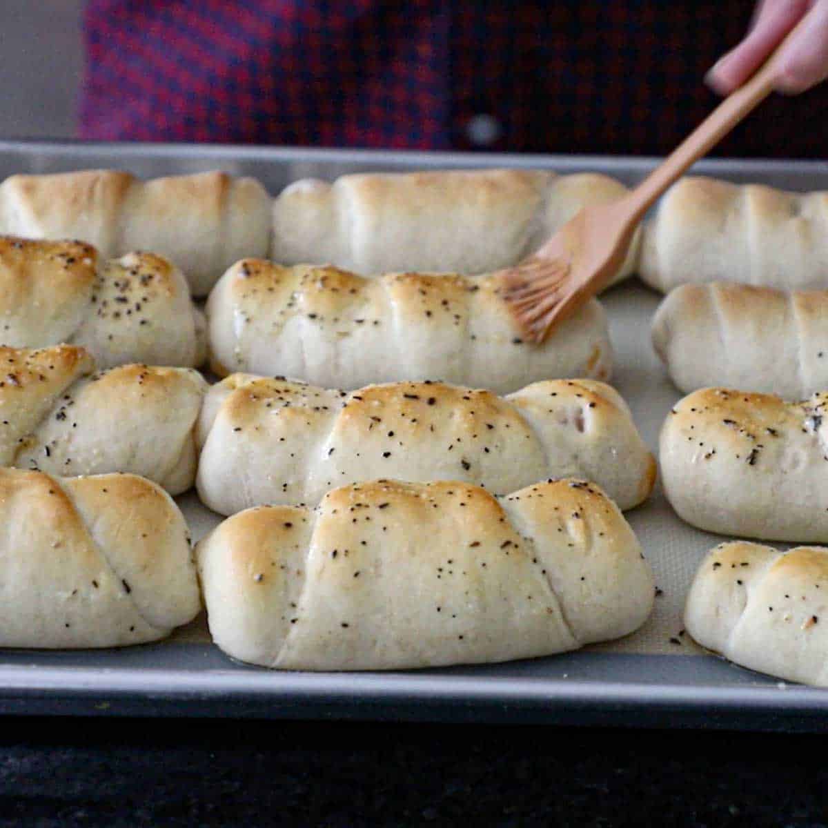 A person using a pastry brush to add seasoned melted butter over the toops of freshly baked Melt-In-Your-Mouth Pepperoni Rolls.