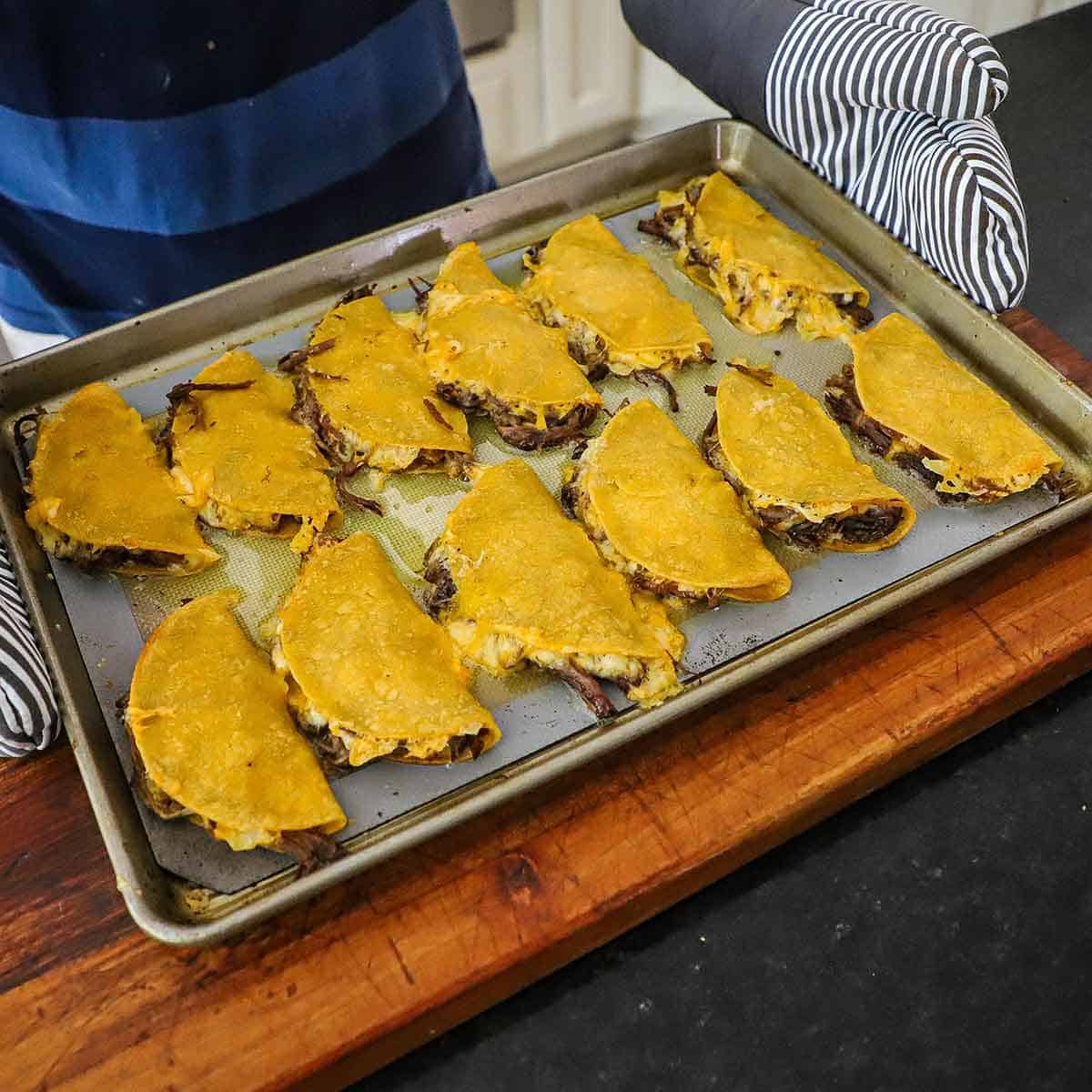 A person wearing oven mitts to hold a baking pan that is filled with freshly baked brisket tacos.