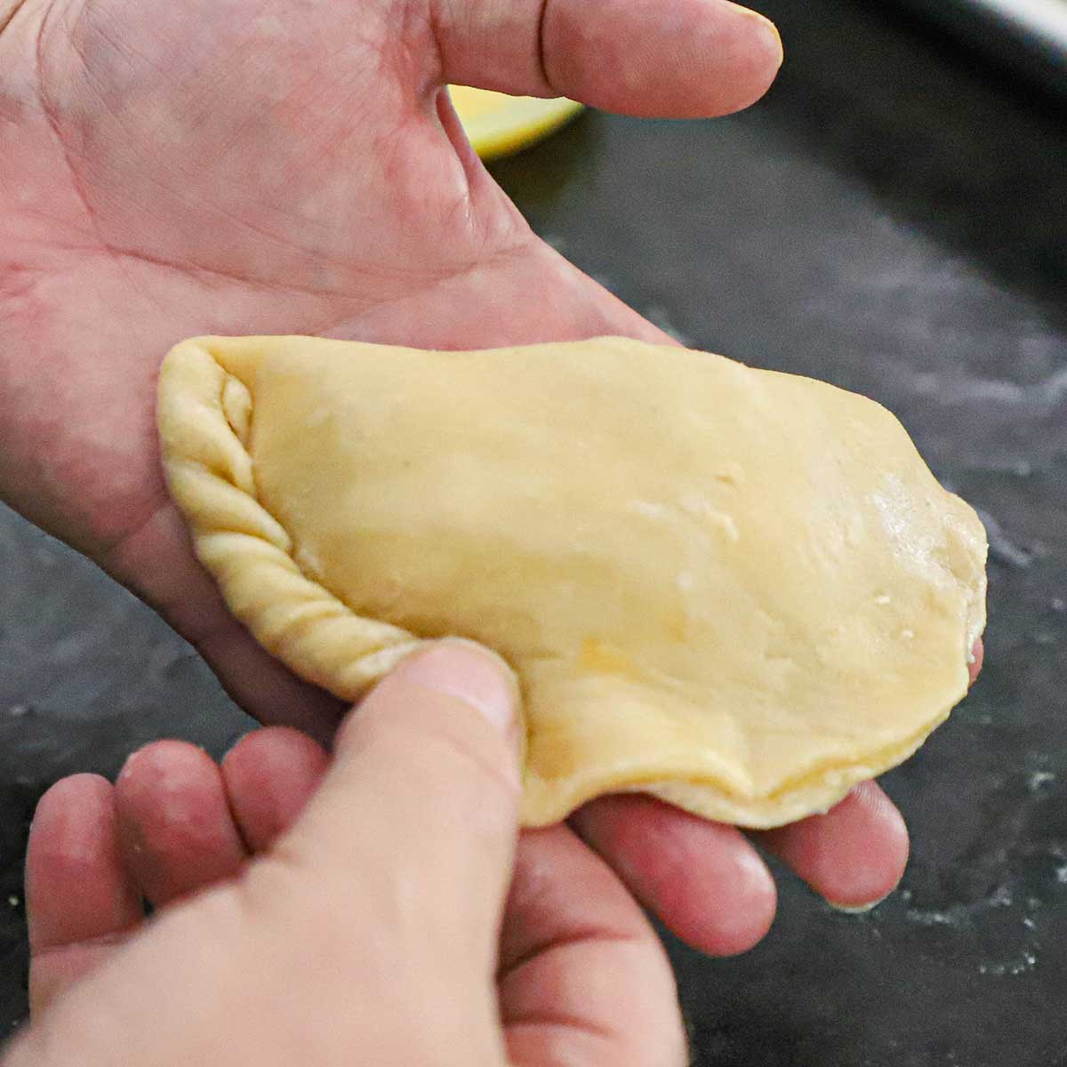 A person using two hands to crimp the edges of an uncooked Argentine Beef Empanada.