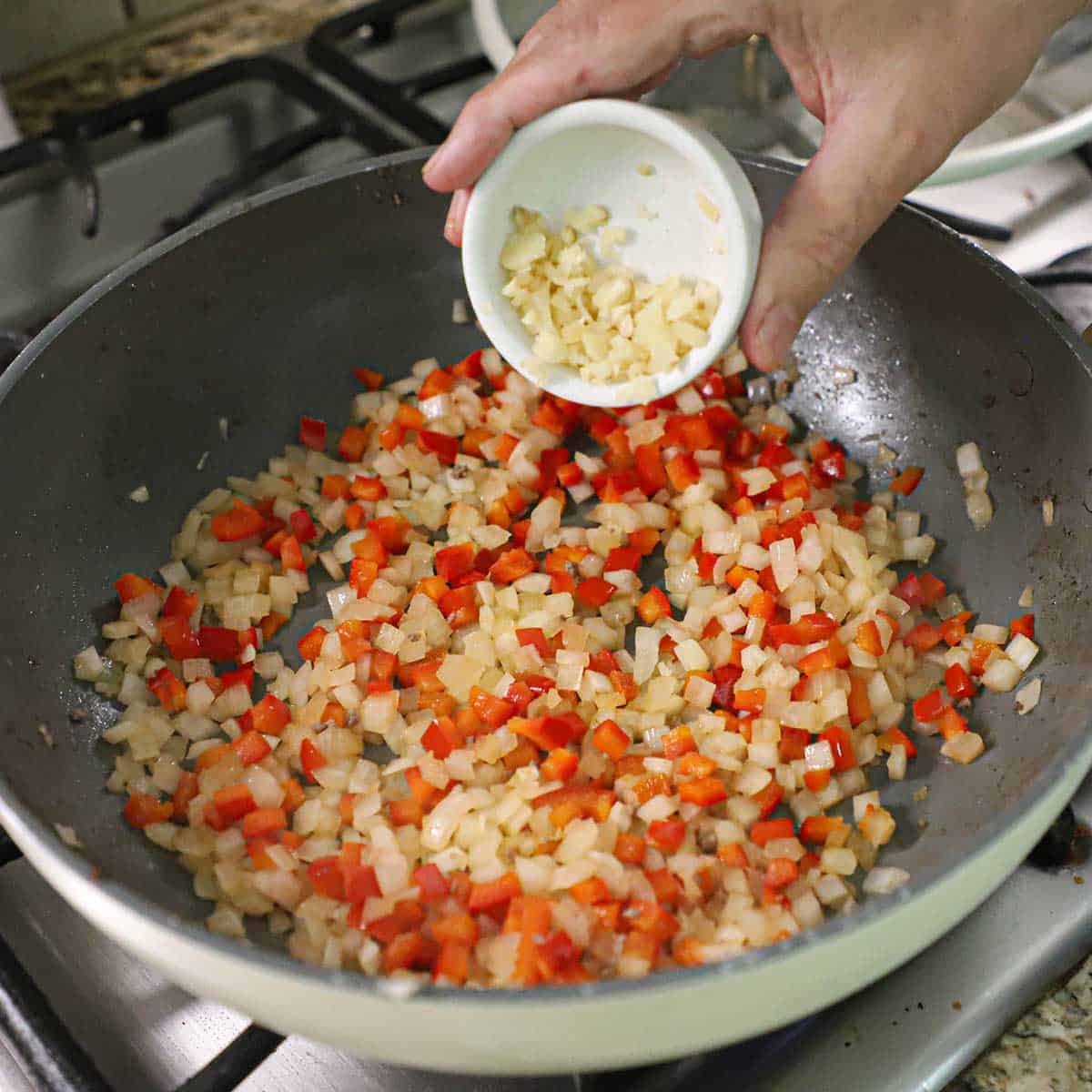A person transferring minced garlic from a small bowl into a large skillet filled with sautéed chopped onions and red bell pepper.