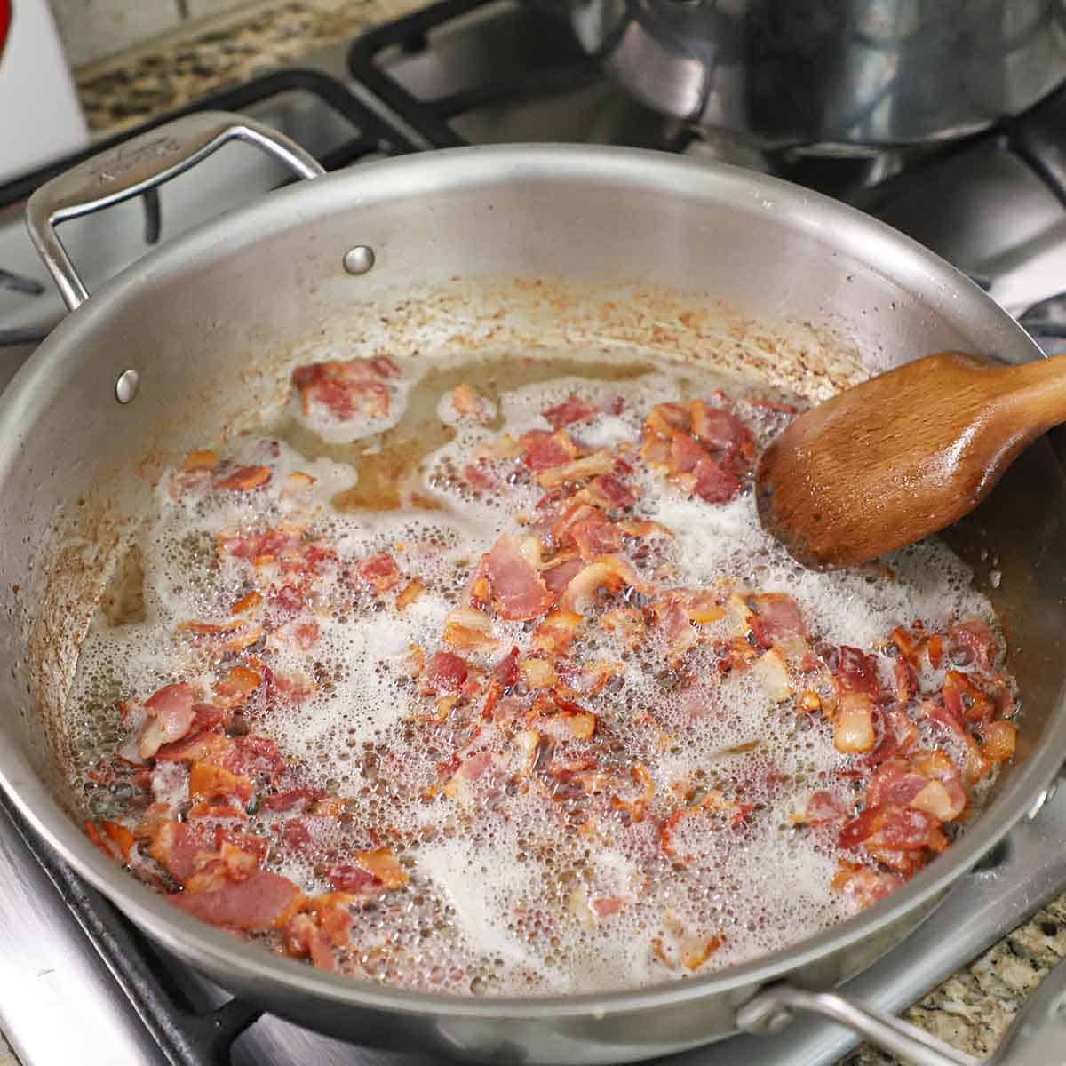 A person using a wooden spoon to stir cooking pieces of bacon in a large stainless steel skillet on a gas stove.