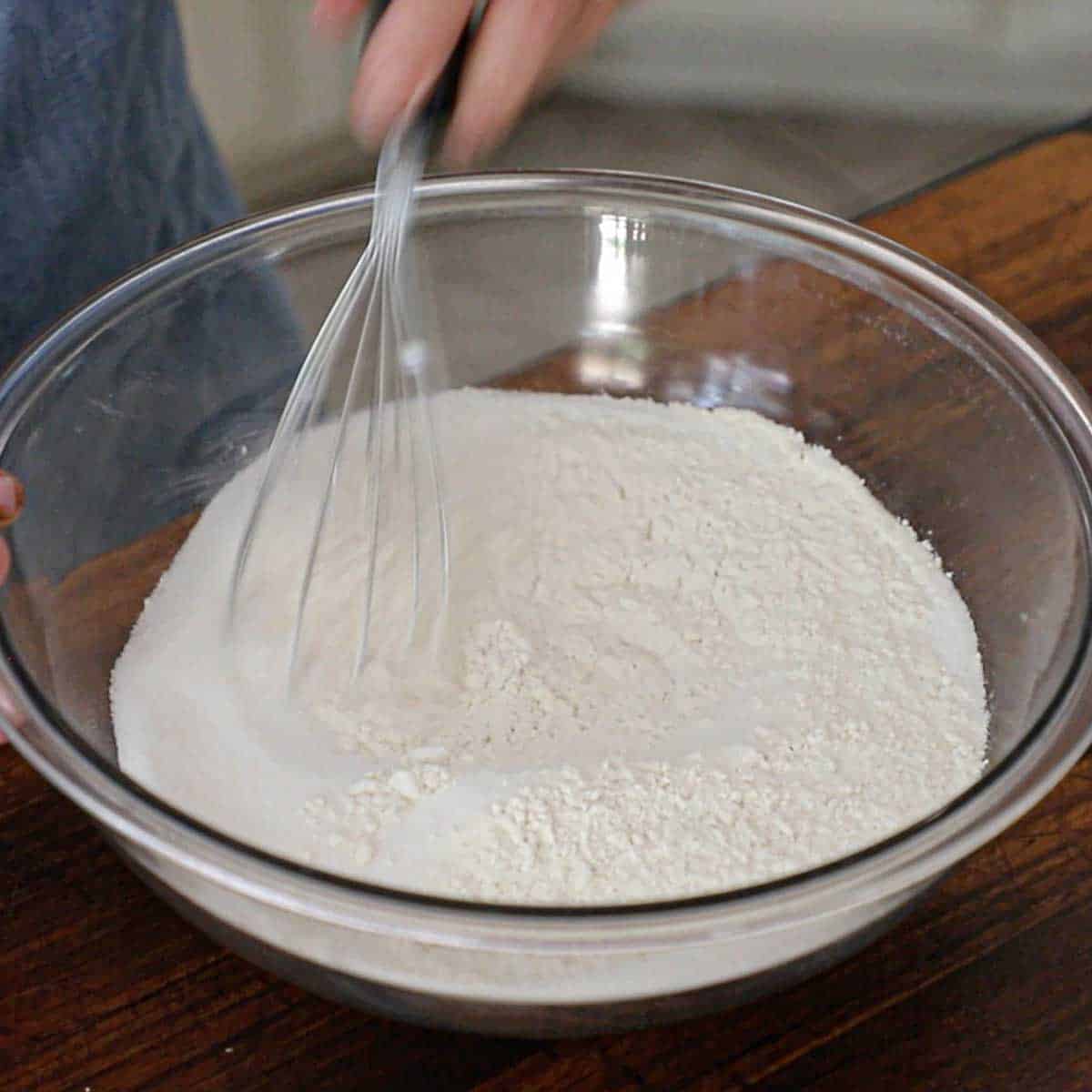 A person using a whisk to combine flour with sugar, baking soda, and salt in a large glass bowl.