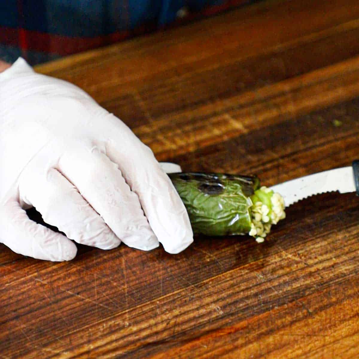 A person wearing protective gloves using his two hands to hold a roasted jalapeño pepper and use a seeder to remove the seeds from the inside of the pepper.
