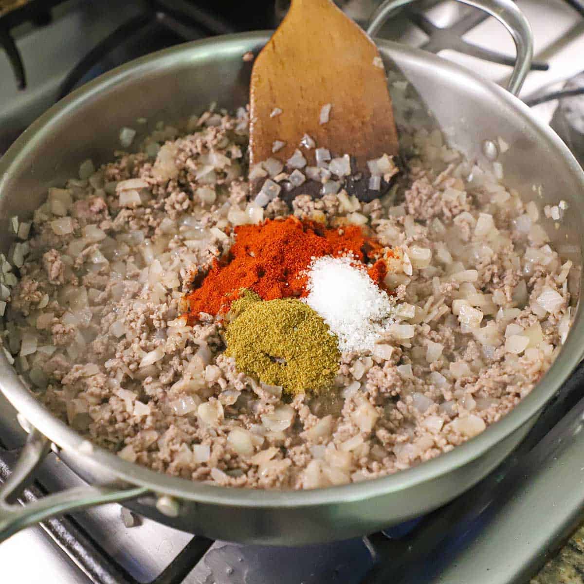 A person using a wooden spatula to stir cooked ground beef, sautéed onions, with smoked paprika, ground cumin, and salt in a large skillet on a gas stove.