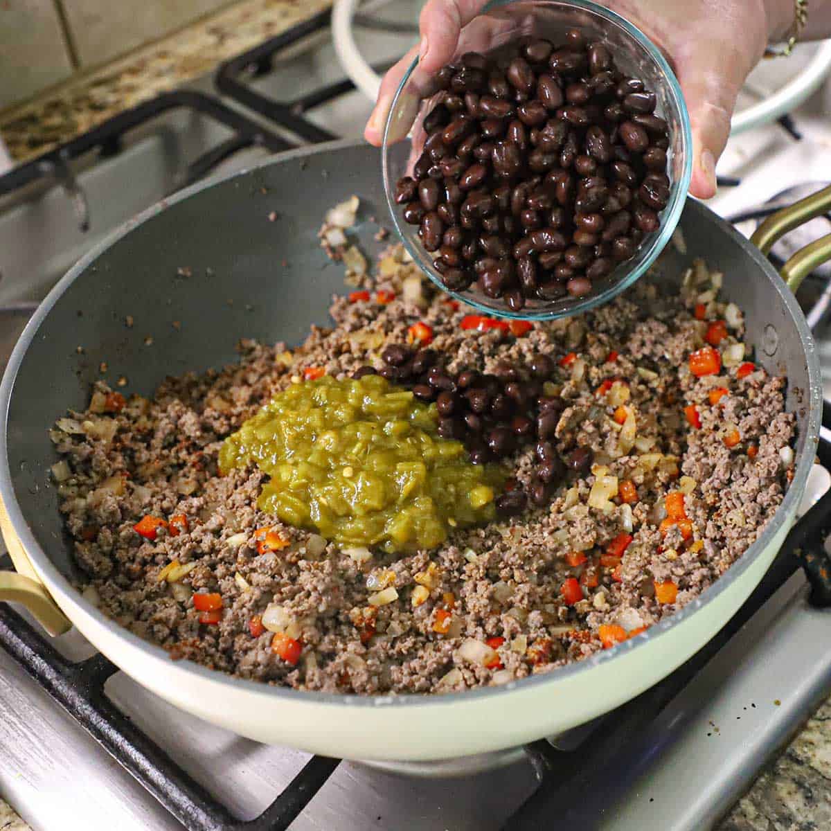 A person transferring black beans from a glass bowl into a skillet that is filled with cooked ground beef, sautéd onions and peppers, and diced green chiles.