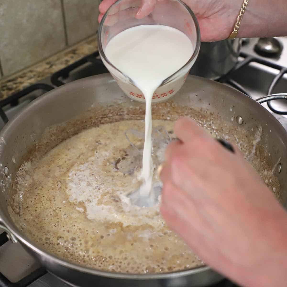 A pers pouring heavy cream from a glass measuring cup into a skillet that he is whisking into a roux in a large skillet.