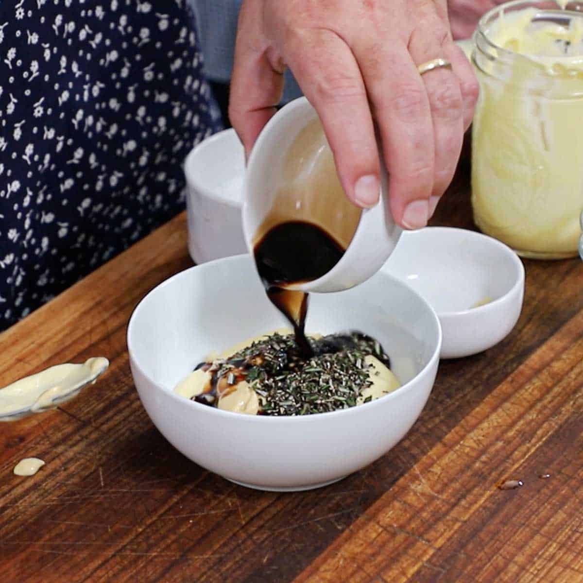 A person pouring balsamic vinegar from a small white bowl into a large white bowl filled with homemade mayonnaise, chopped rosemary, and ground black pepper.