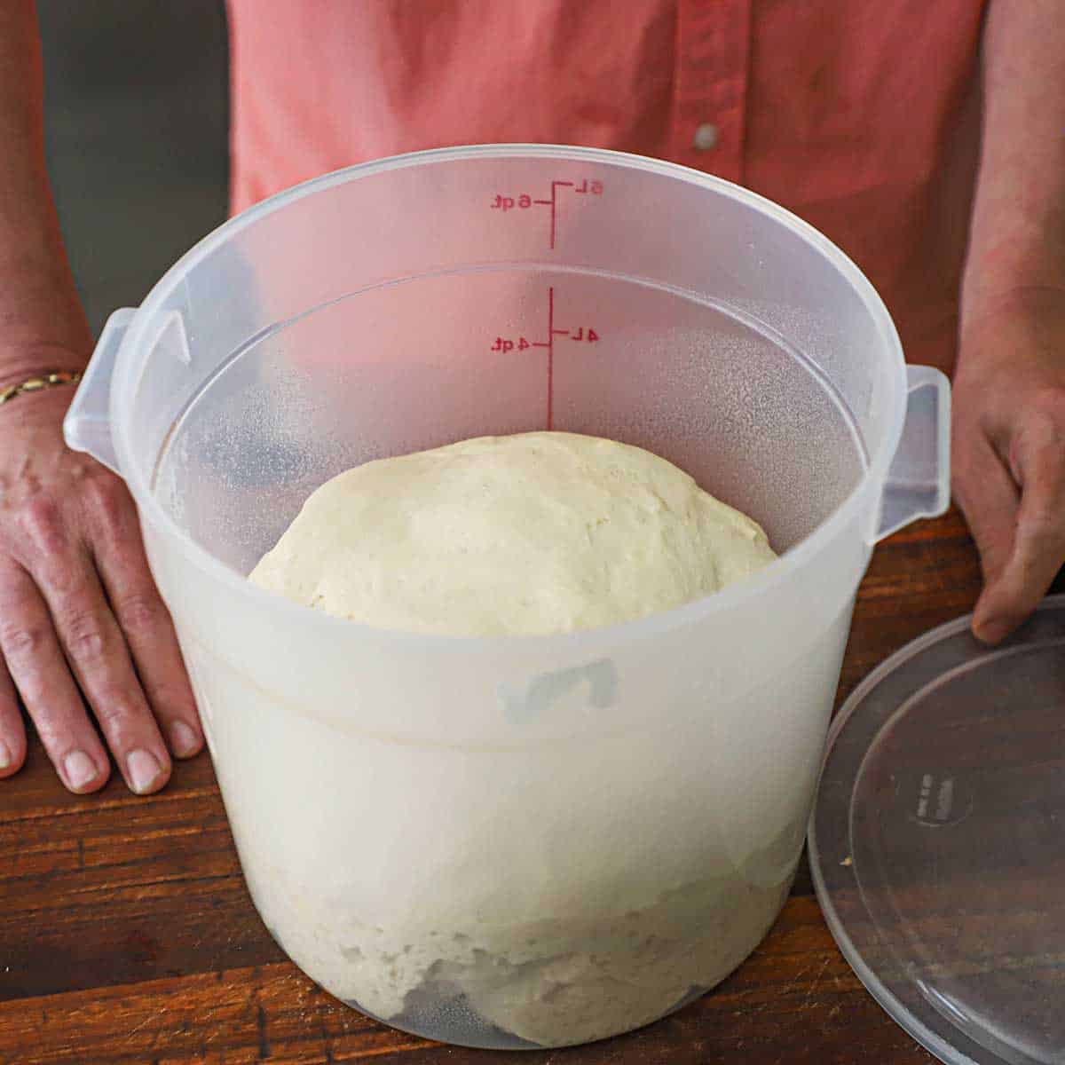 A person standing behind a proofing container that is holding a large all of pizza dough that is large and nearly halfway filling up the container.