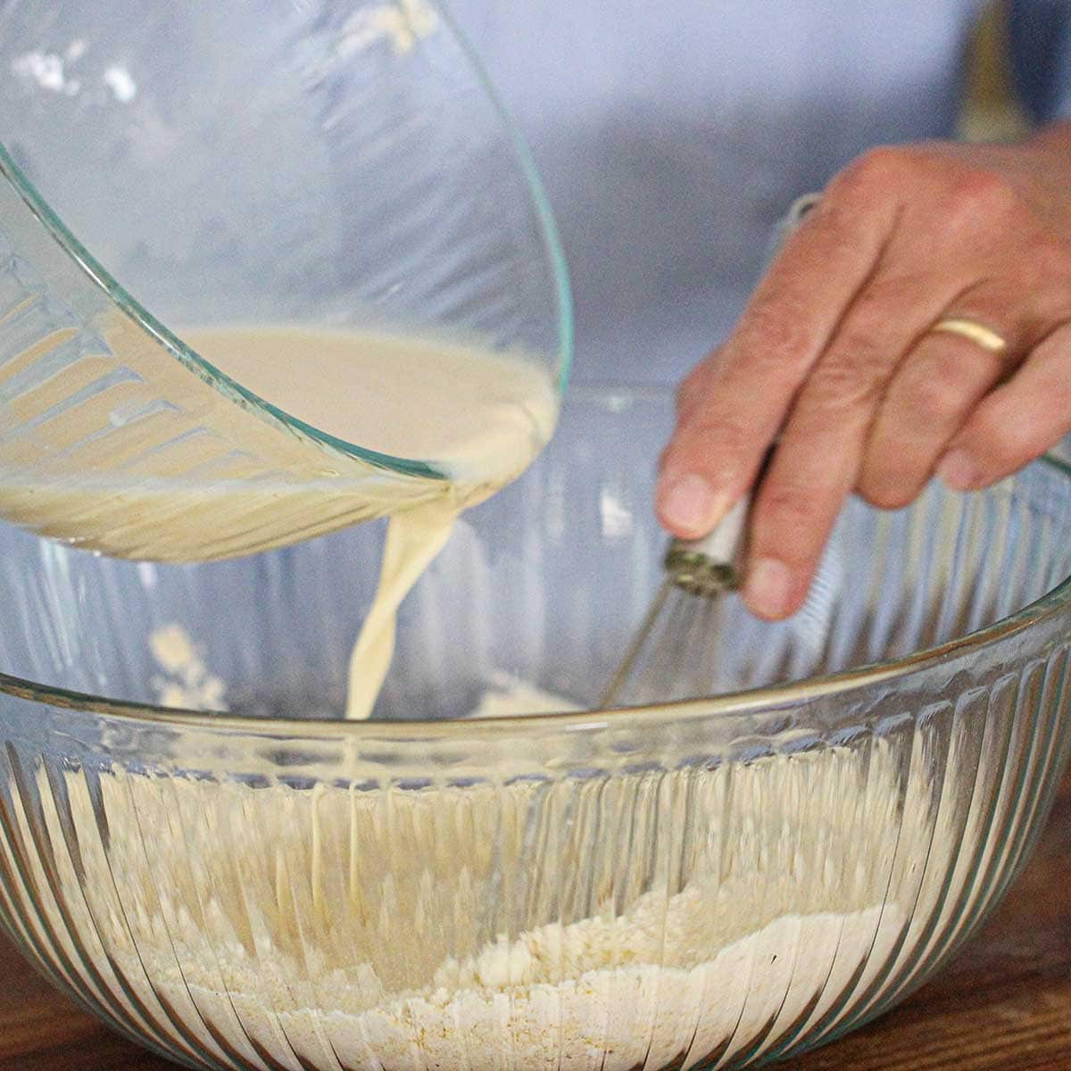 A person pouring a mixture of buttermilk, eggs, and hot sauce from a small glass bowl into another bowl filled with the dry ingredients for Southern Hush Puppies.
