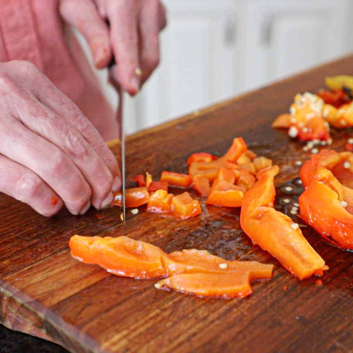 A person using a knife to cut a roasted bell pepper into bite-sized pieces on a wooden cutting board.