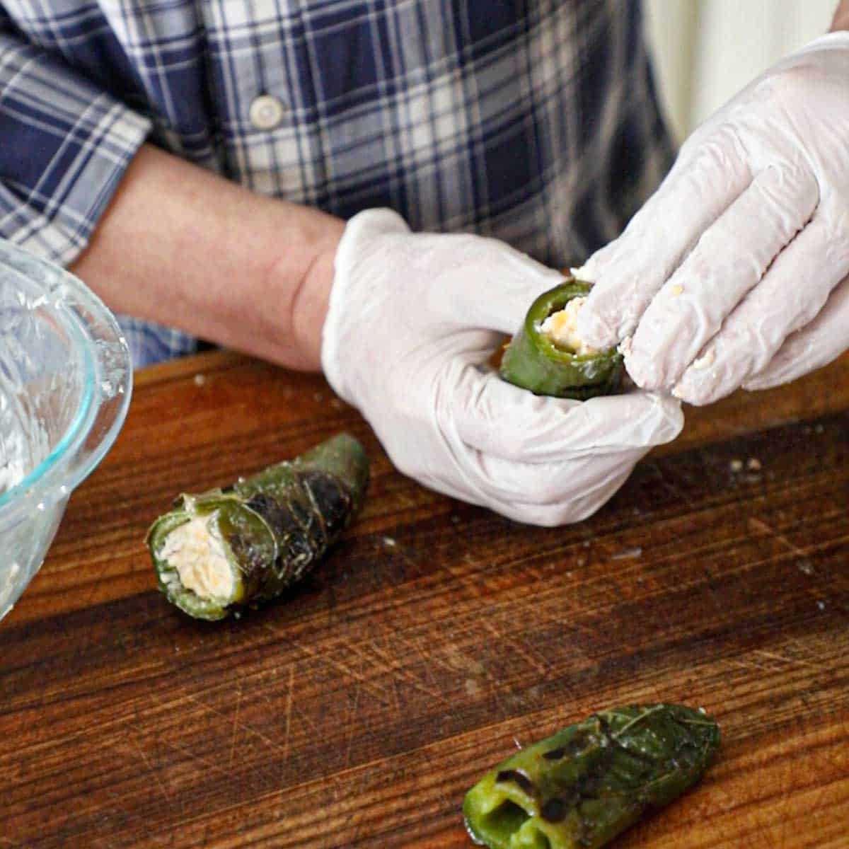 A person wearing protective gloves stuffing softened cheese into the cavity of a seeded roasted jalapeño pepper.