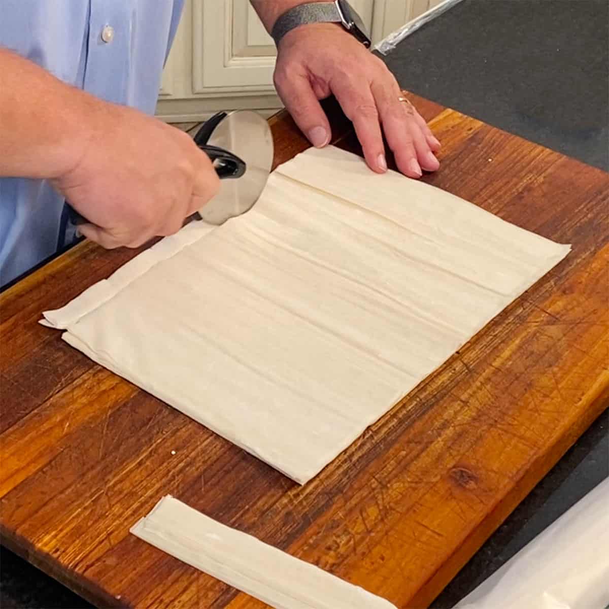 A person using a pizza cutter to trim down layers of phyllo dough on a wooden cutting board.