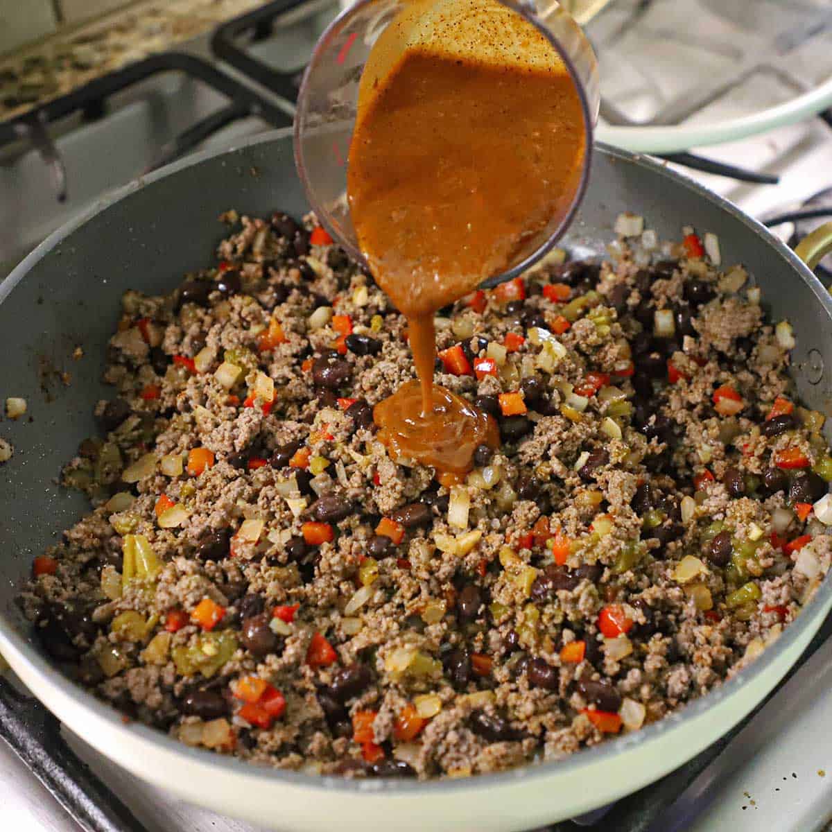 A person pouring red enchilada sauce from a measuring cup into a large skillet filled with a Mexican ground beef and black bean mixture.