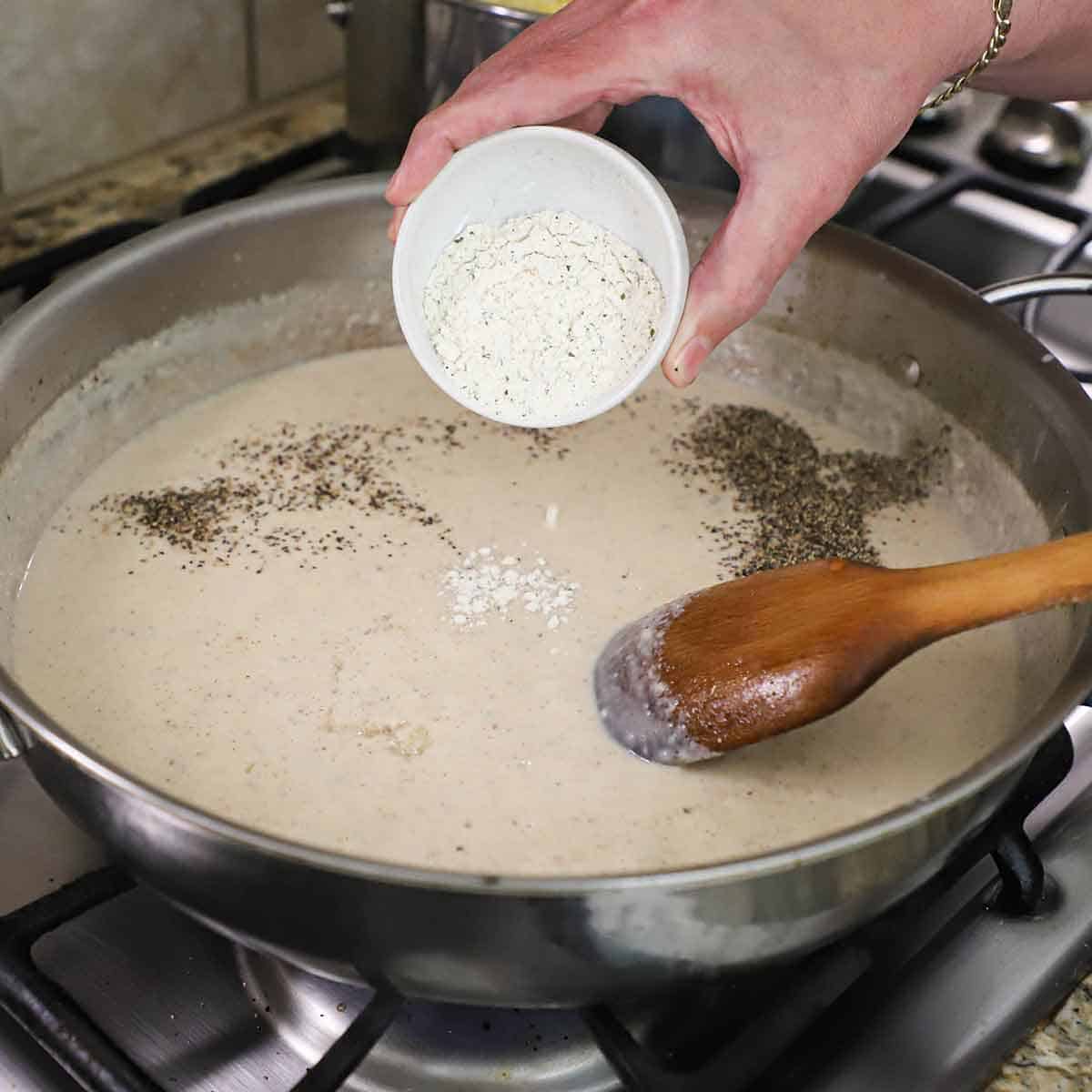 A person transferring Ranch seasoning from a small bowl into a large skillet filled with a bechamel sauce with black pepper also sprinkled on the surface.