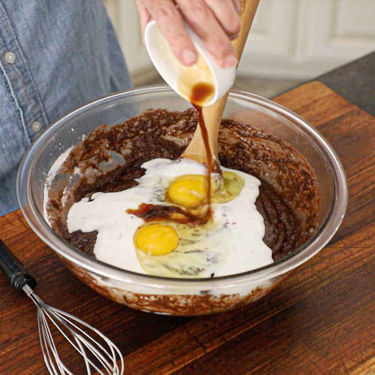 A person pouring vanilla extract from a small white bowl into a glass bowl filled with a chocolate cake batter that is topped with buttermilk and two whole eggs.