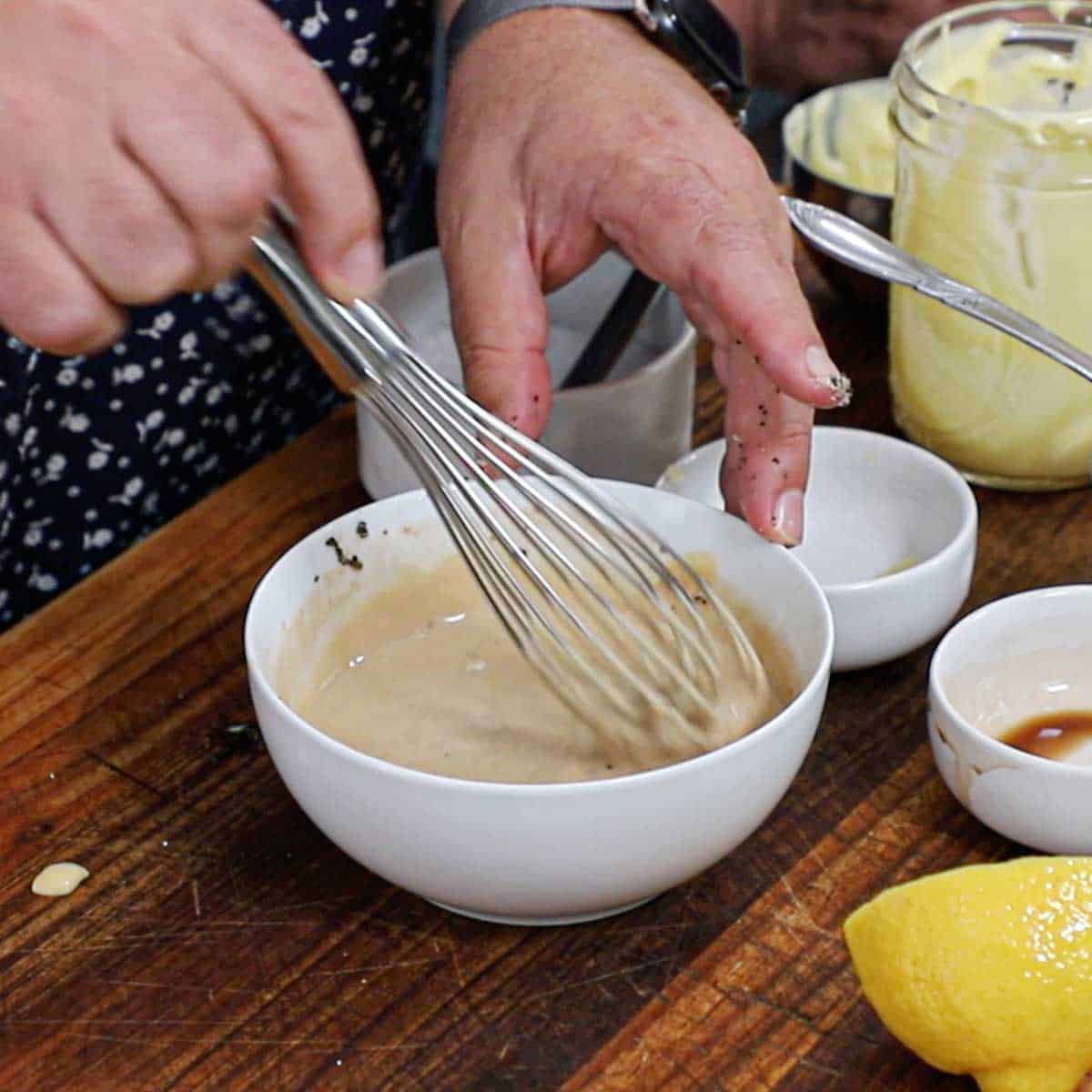 A person using a whisk to mix the components of rosemary garlic aioli in a white bowl on a wooden cutting board.