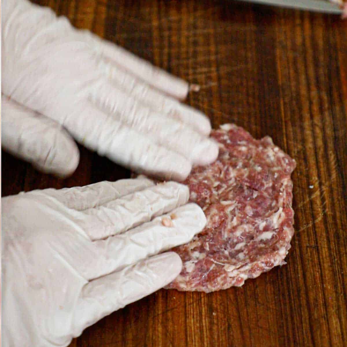 A person using his two hands to flatten Italian sausage into a rectangle on a wooden cutting board.