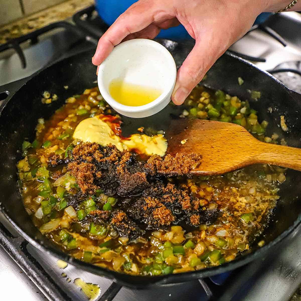 A person pouring a small amount of apple cider vinegar into a skillet filled with simmering chopped onions and peppers, and brown sugar, molasses, Dijon mustard, and BBQ sauce.