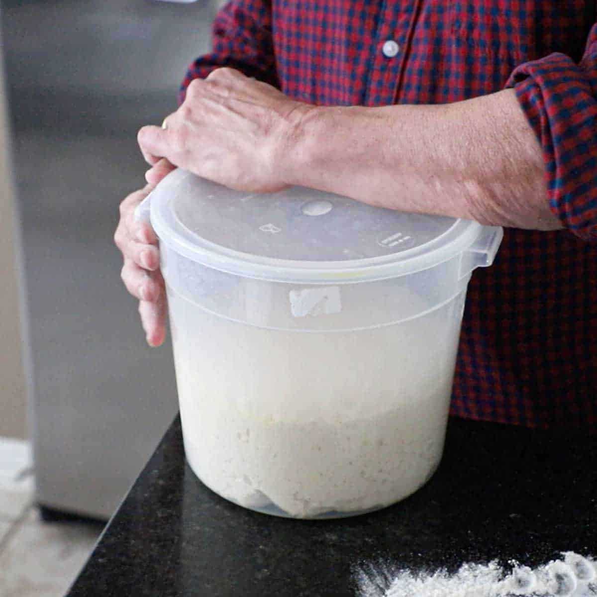 A person using his hand to remove the lid from a plastic proofing container that is filled with dough that has been proofed and doubled in size.