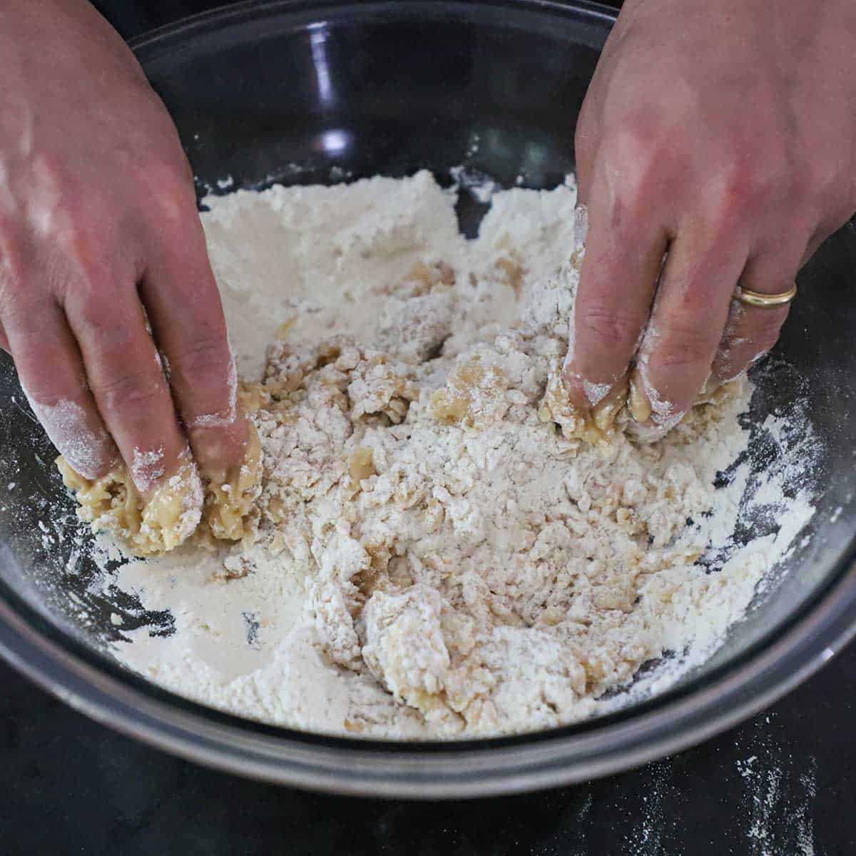 A person using his two hands to work softened pork lard into flour in a large glass bowl.