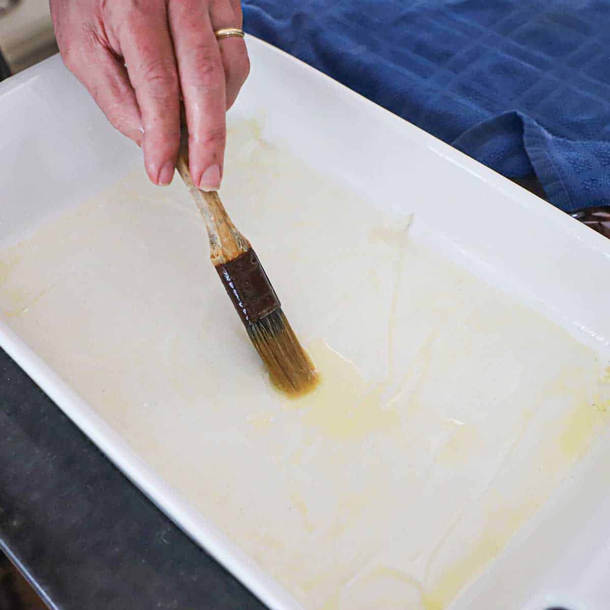 A person using a pastry brush to brush melted butter over the bottom and sides of a 9x13" baking dish.