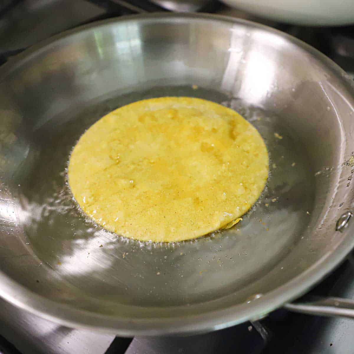 A corn tortilla lightly being flash fried in a small silver skillet filled with a layer of hot oil on a gas stove.