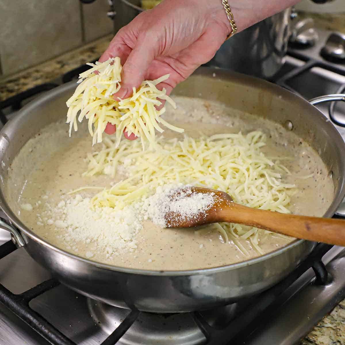 A person using his hand to transfer shredded white cheddar cheese into a large skillet filled with a simmering bechamel sauce along with Parmesan cheese sprinkled on top.