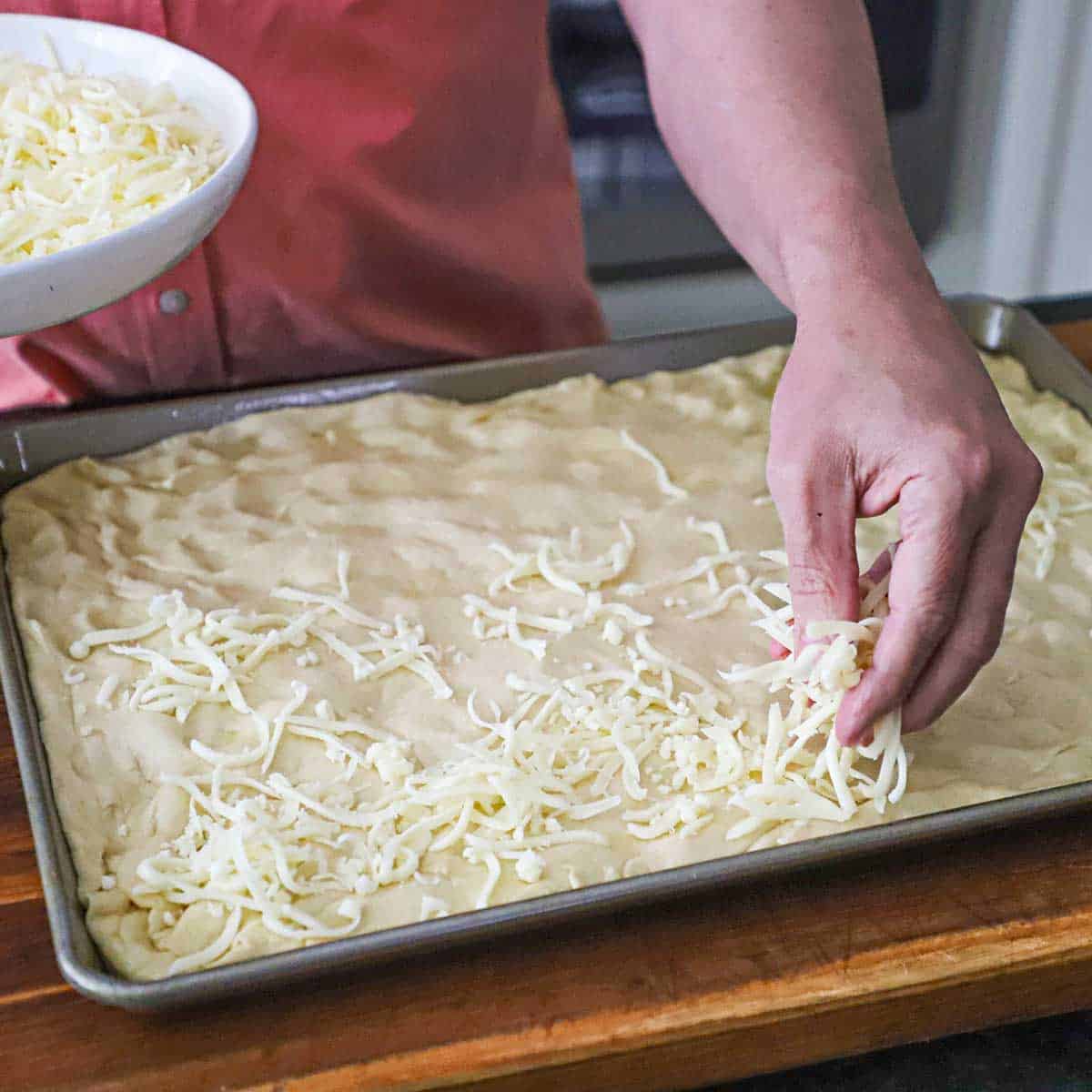 A person sprinkling shredded mozzarella over the top of pizza dough that has been stretched to the inside edges of a half sheet pan.