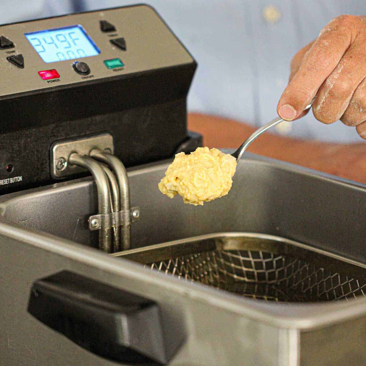 A person getting ready to lower a ball of hush puppies batter from a spoon into a deep fryer filled with hot oil.