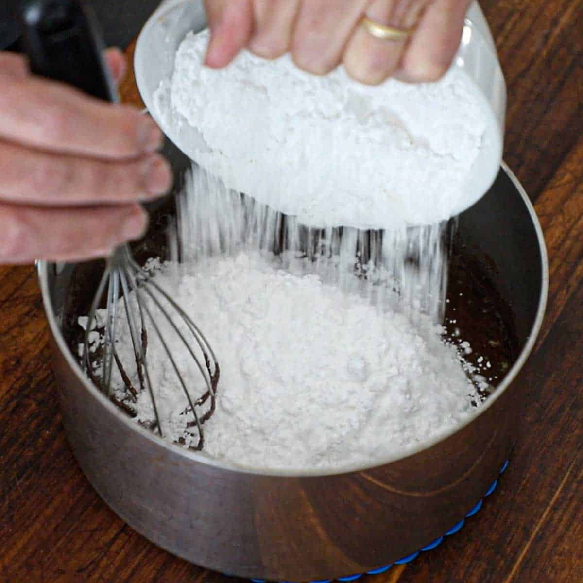 A person transferring powdered sugar from a bowl into a saucepan filled with a liquid chocolate, butter, and milk mixture.