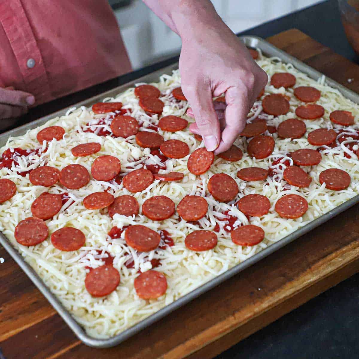 A person placing sliced pepperoni over the top of a half sheet pan that is filled with pizza dough and topped with pizza sauce, shredded mozzarella, and more pepperoni.