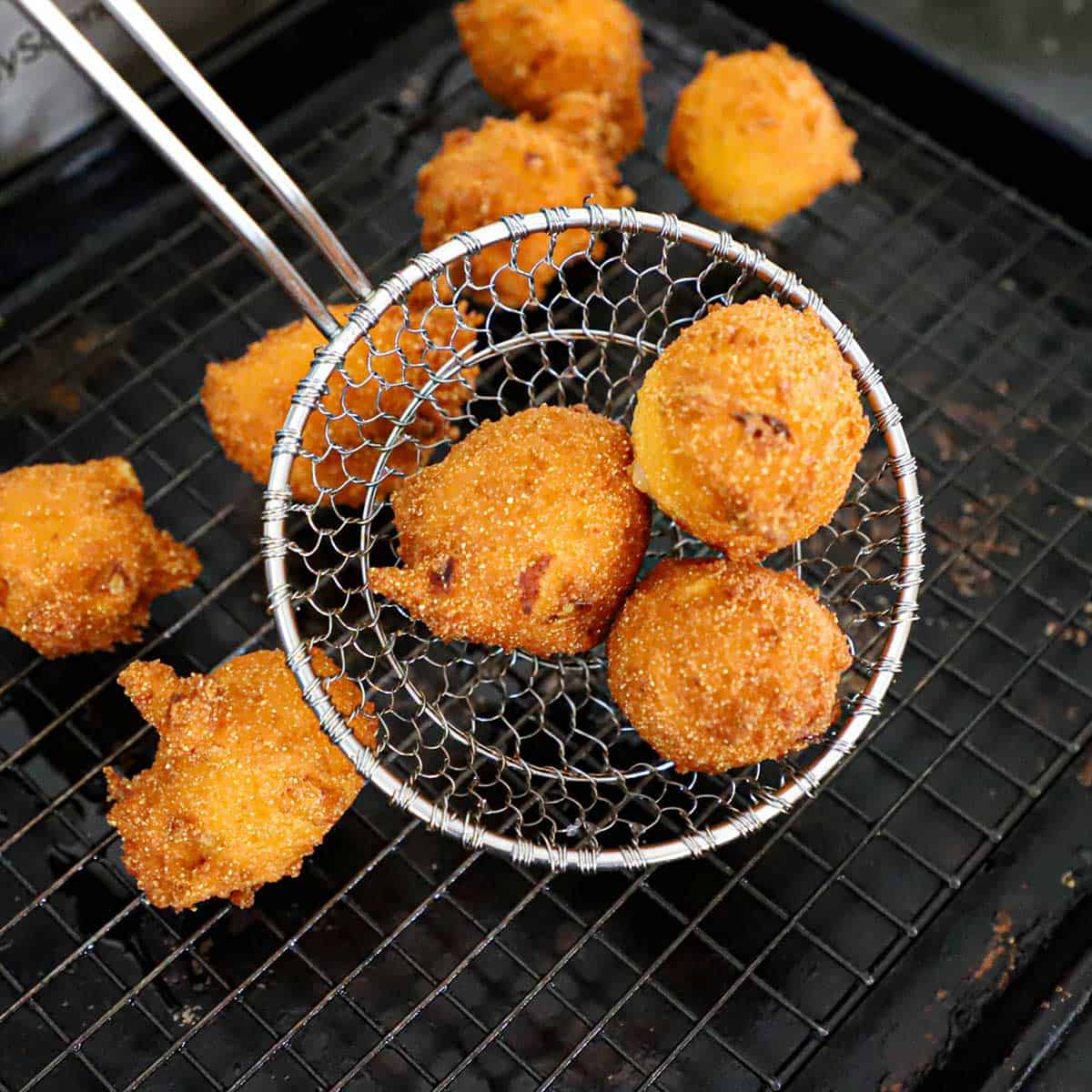 A person using a metal spider to hold three freshly fried Southern Hush Puppies over a baking pan lined with a baking rack.