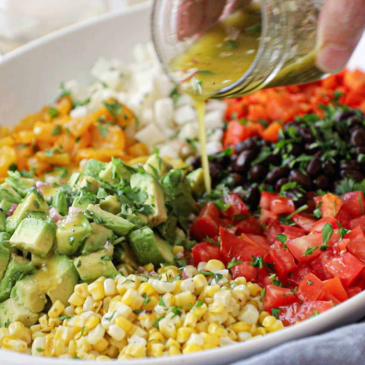 A person pouring a citrus-honey vinaigrette from a small jar over the tops of a deconstructed Southwestern Summer Salad in a large white platter.