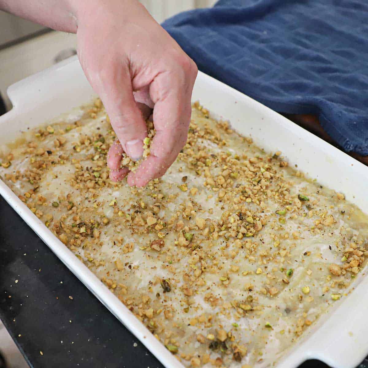 A person sprinkling a mixture of spiced pieces of walnuts and pistachios over stacks of buttered sheets of phyllo dough in a 9x13" baking dish.