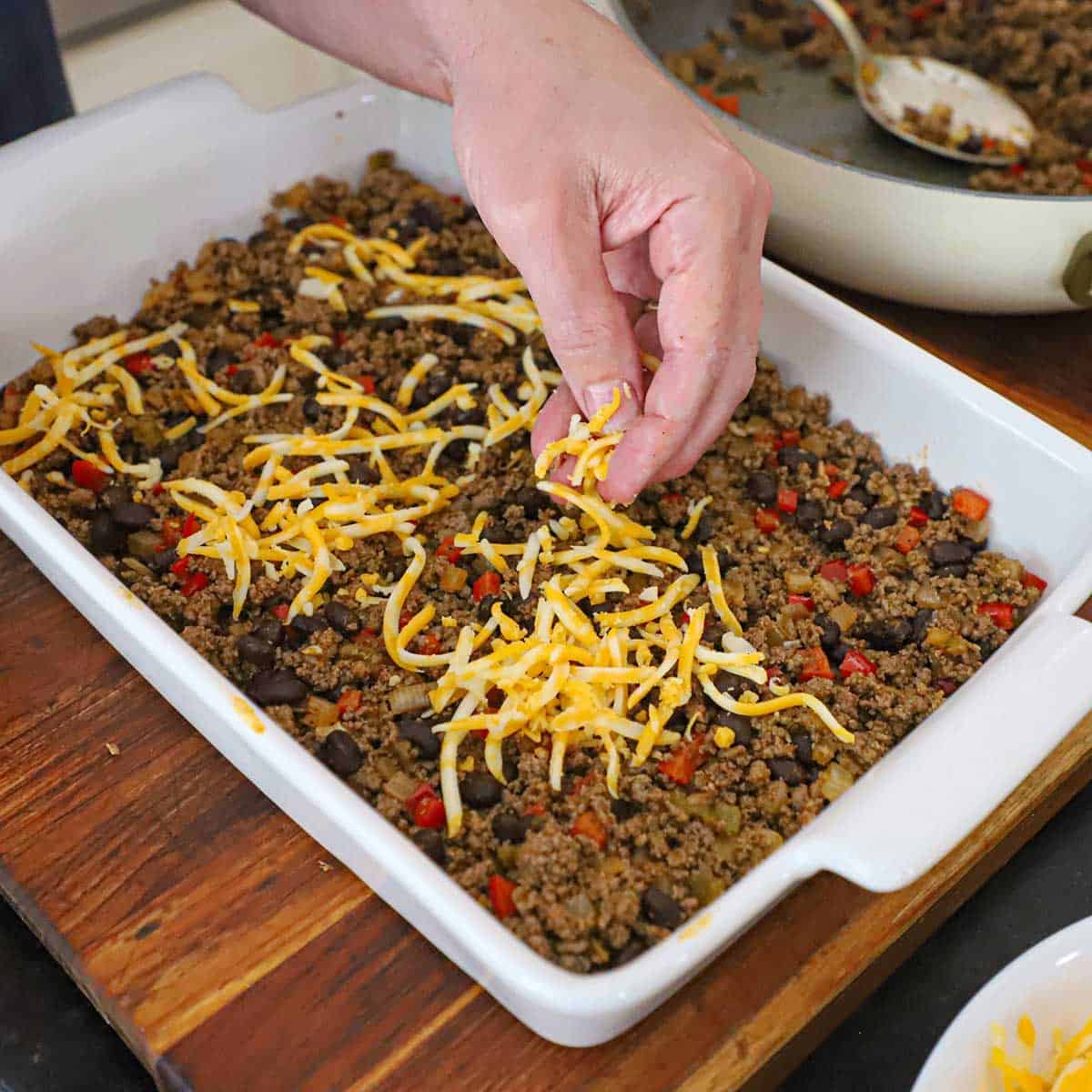 A person sprinkling shredded Colby Jack cheese over the top of Mexican ground beef and black bean mixture that has been spread out over corn tortillas in a 9 by 13-inch baking dish.