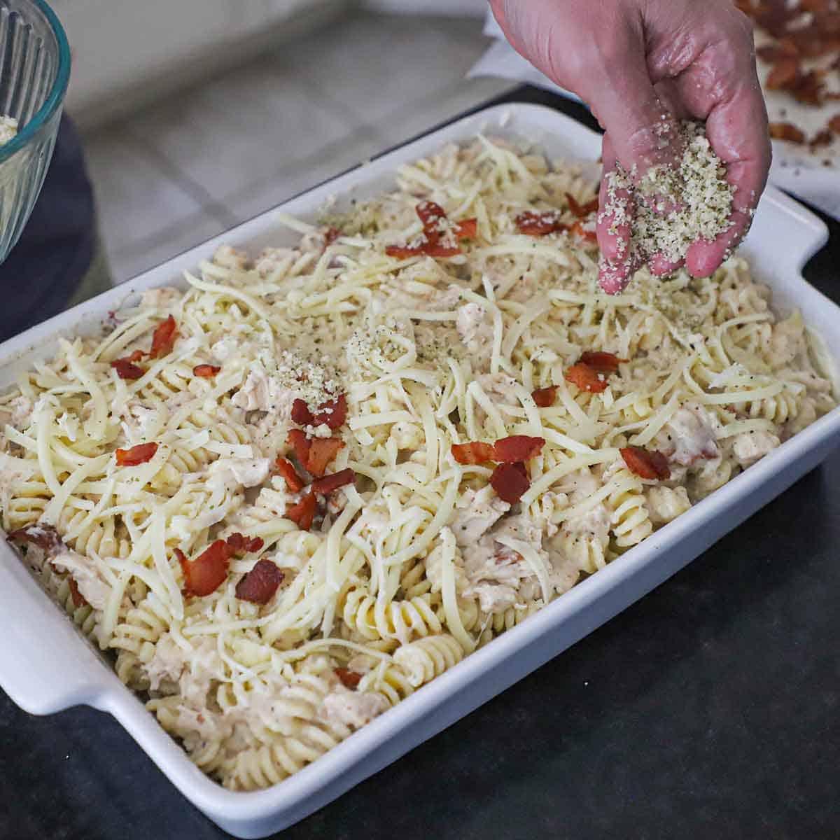 A person sprinkling butter Panko breadcrumbs over the top of an unbaked chicken bacon ranch casserole in a white baking dish.