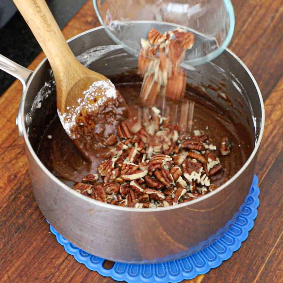 A person transferring chopped pecans into a chocolate frosting mixture in a silver saucepan.