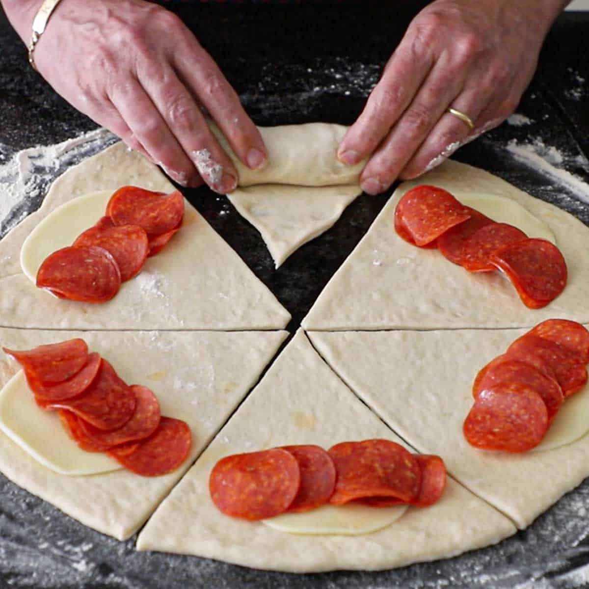 A person using both hands to roll a triangular piece of dough that is filled with Provolone cheese and thinly sliced pepperoni into a pepperoni roll.