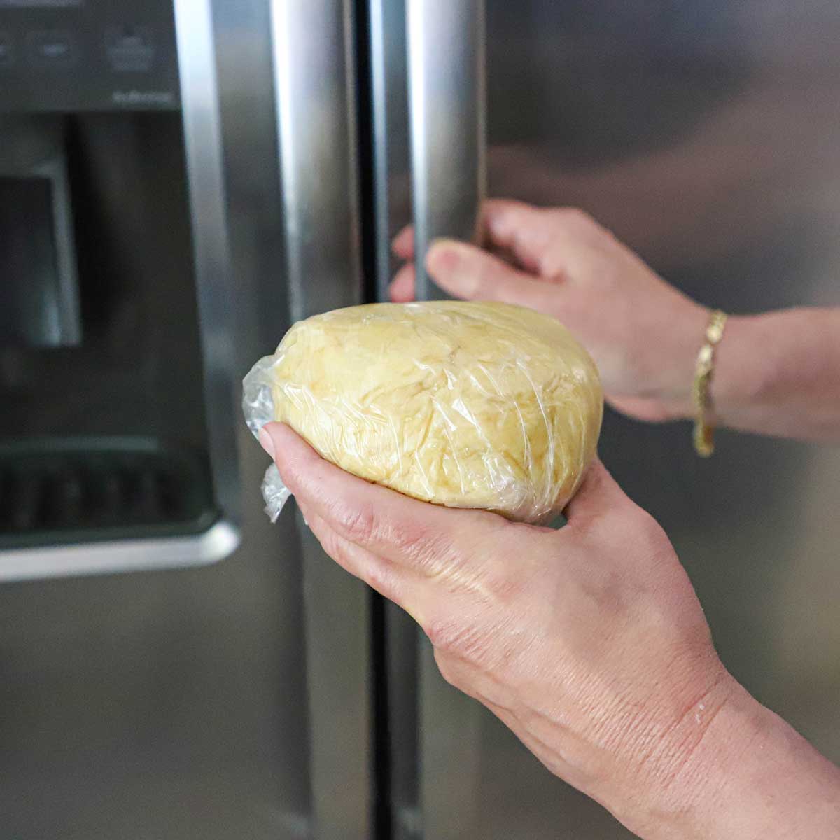 A person holding a ball of empanada dough that is wrapped in plastic wrap that he is about to place in a refrigerator.
