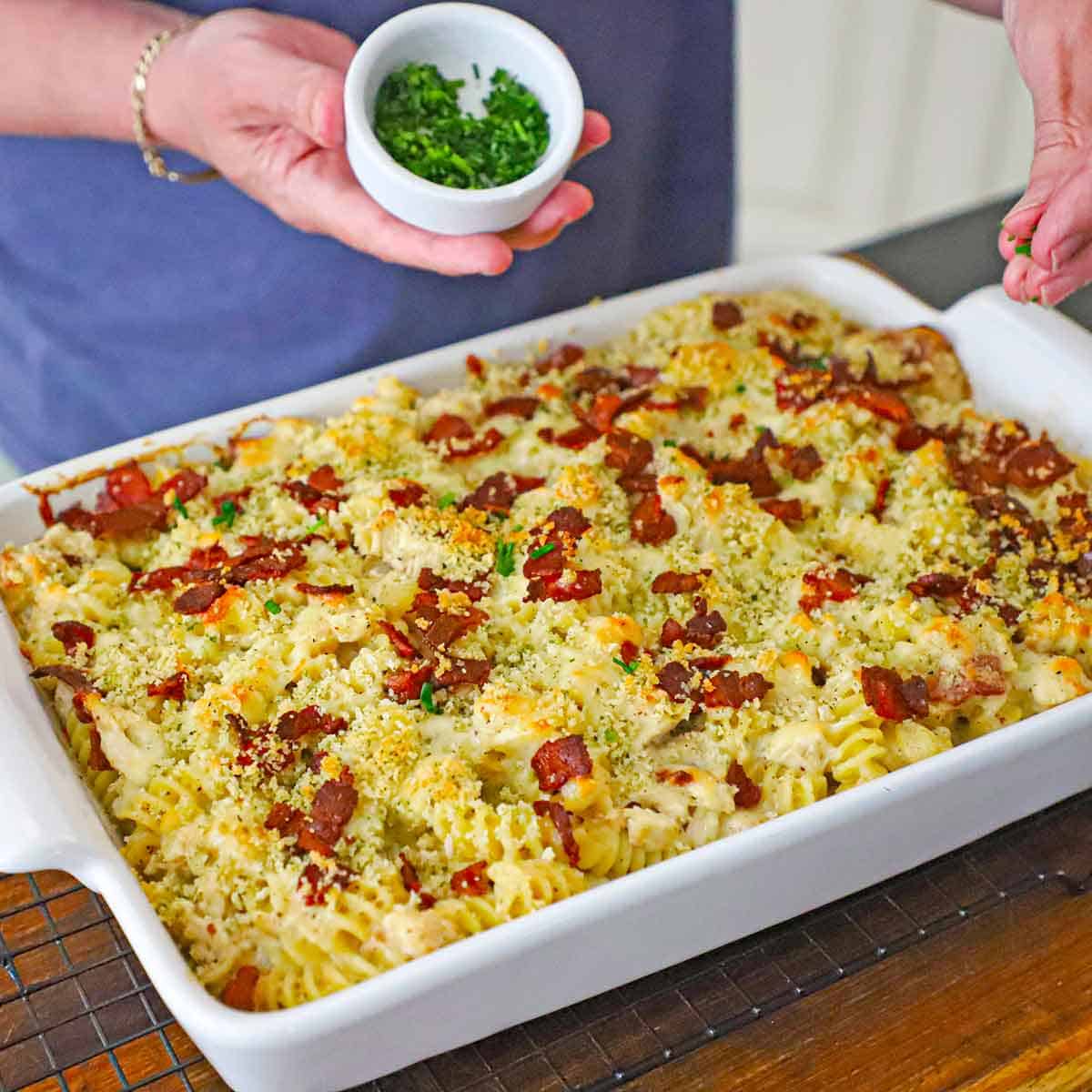 A person sprinkling snipped chives of the top of a freshly baked chicken bacon ranch casserole in a 9 by 13 inch baking dish resting on a baking rack.