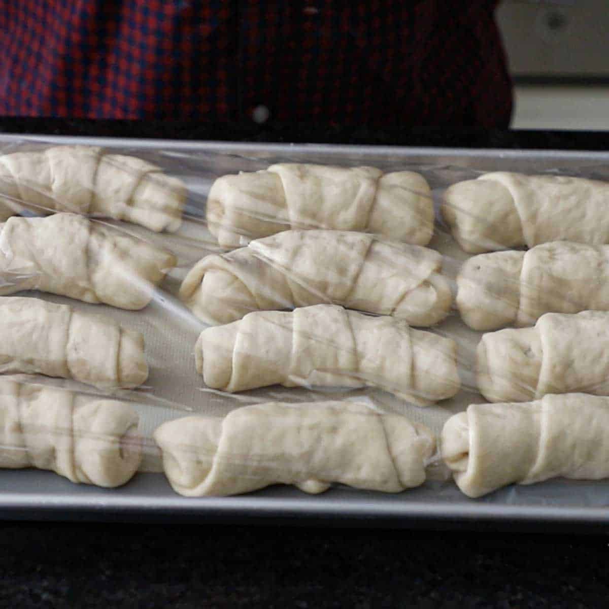 A person placing plastic wrap over a large baking pan that is filled with unbaked Melt-In-Your-Mouth Pepperoni Rolls.