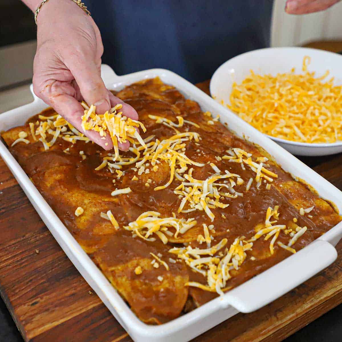 A person sprinkling a top layer of shredded cheese over the top of an uncooked beef enchilada casserole.
